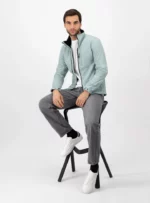 Hombre sentado usando chamarra de color azul claro, pantalones gris y tenis blancos.